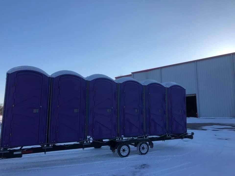 Purple portable toilets on a trailer in a snowy area, a building in the background.