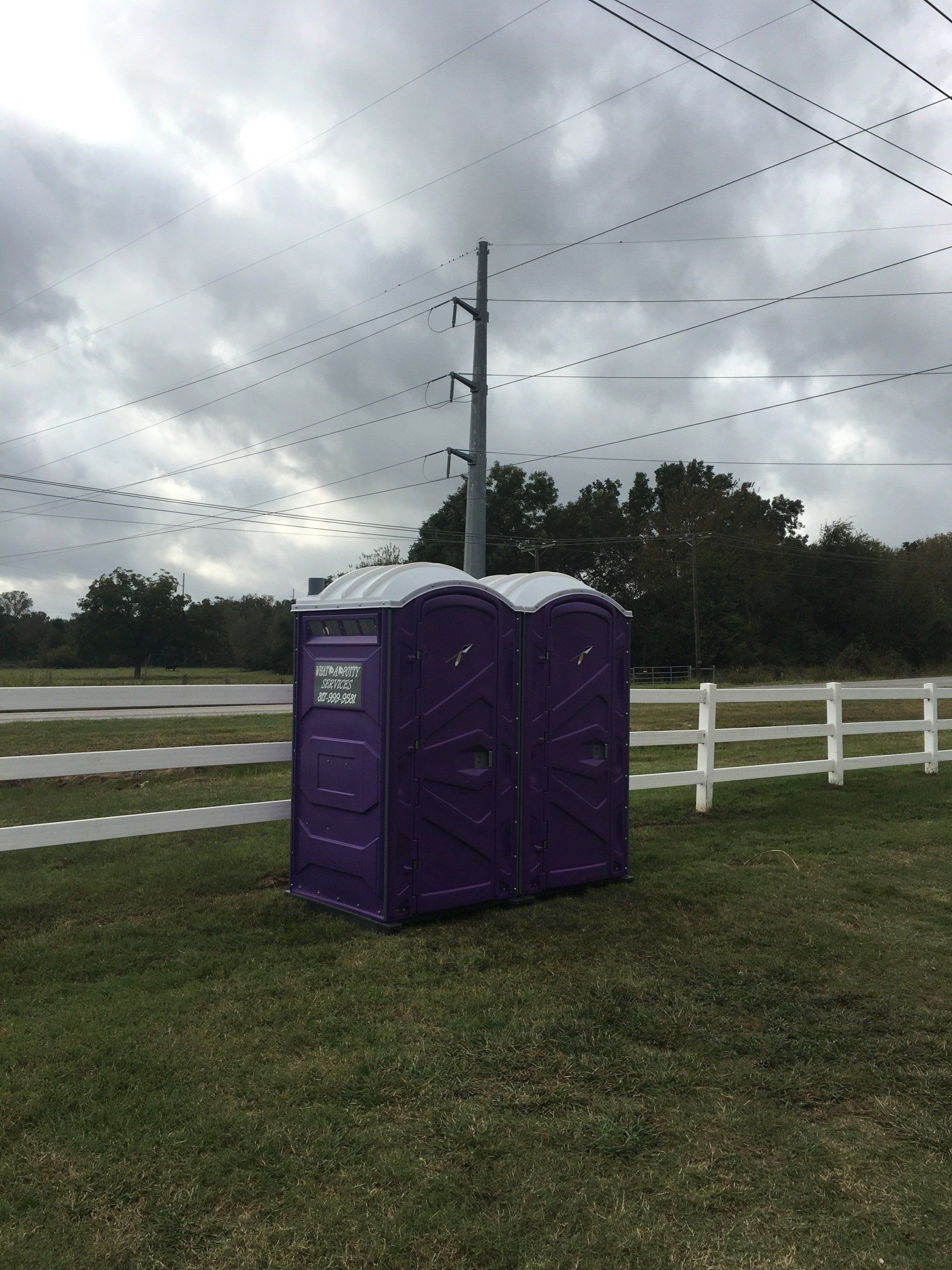 Purple portable toilets on grass with a white fence and power lines against a cloudy sky.