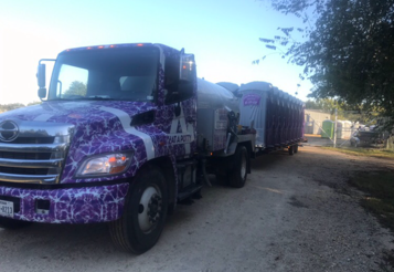 A purple and white wrapped truck pulling a trailer of portable toilets on a dirt road.