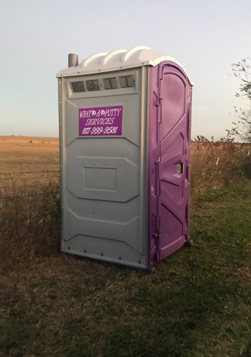 Portable restroom in a field, gray with purple door, company logo, under a blue sky.