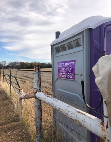 Purple and grey portable toilet next to a rusty fence in a field under a cloudy sky.