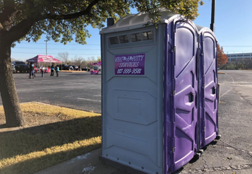 Two purple portable toilets in a parking lot, with people and a pink tent visible in the background.