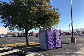 Two purple porta-potties sit in a parking lot next to a tree, with cars and a tent visible in the background.
