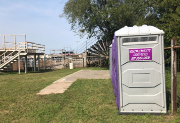 Portable toilet in front of a rodeo arena. White and purple. Outdoors, grass, and a sunny sky.