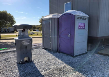 Three portable toilets and a handwashing station on a gravel lot next to a building on a sunny day.