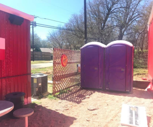 Two purple portable toilets next to a red building with a lattice fence in an outdoor setting.