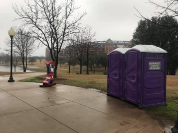 Two purple portable toilets and a red boot sculpture on a walkway near a park and stadium under overcast skies.