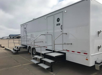 White portable restroom trailer with steps and handrails on a paved surface.