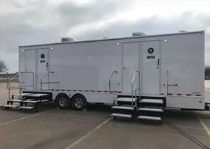 A white restroom trailer with stairs attached to it is parked in a parking lot