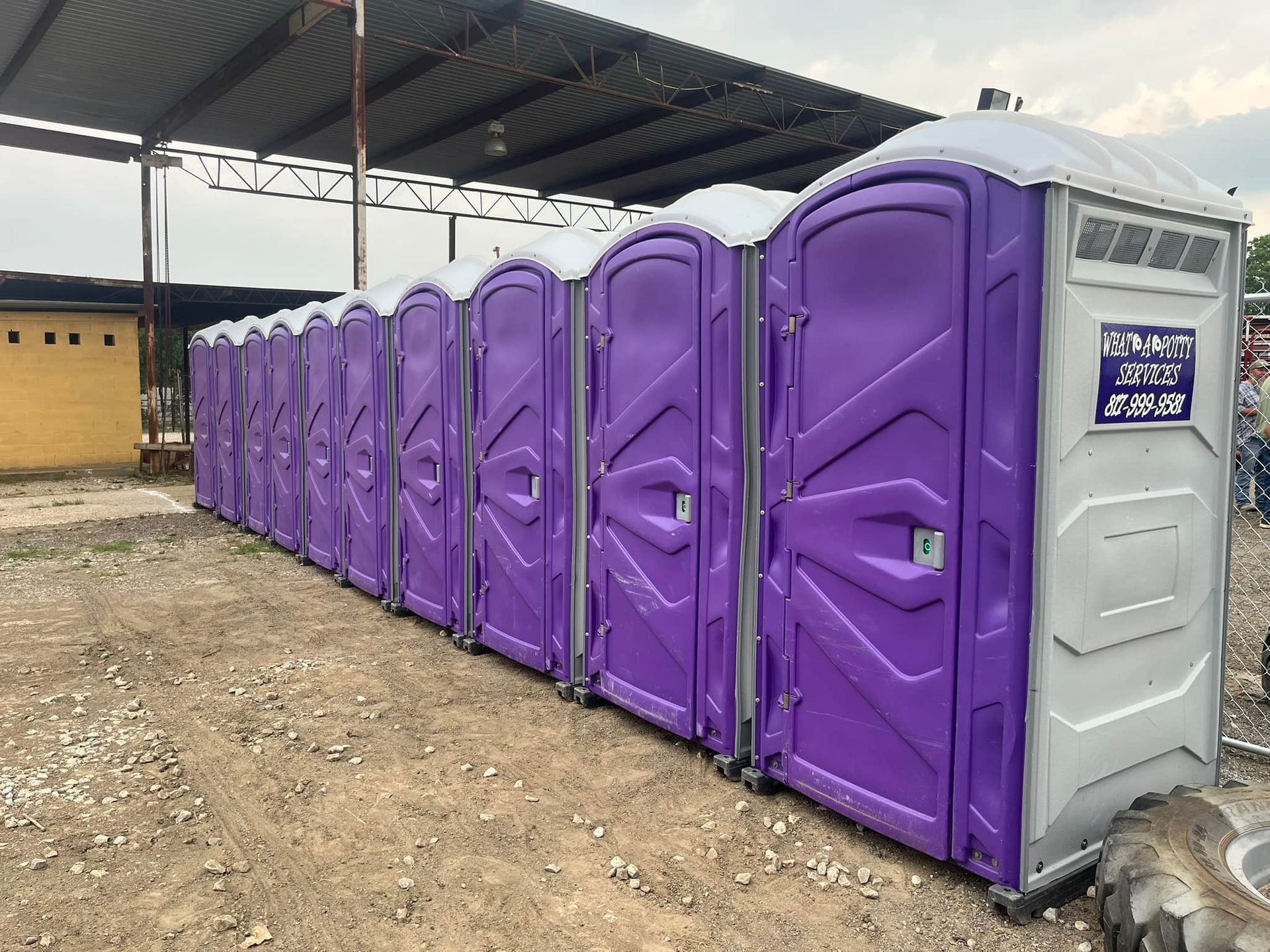 A row of purple portable toilets on a gravel surface under a covered structure.