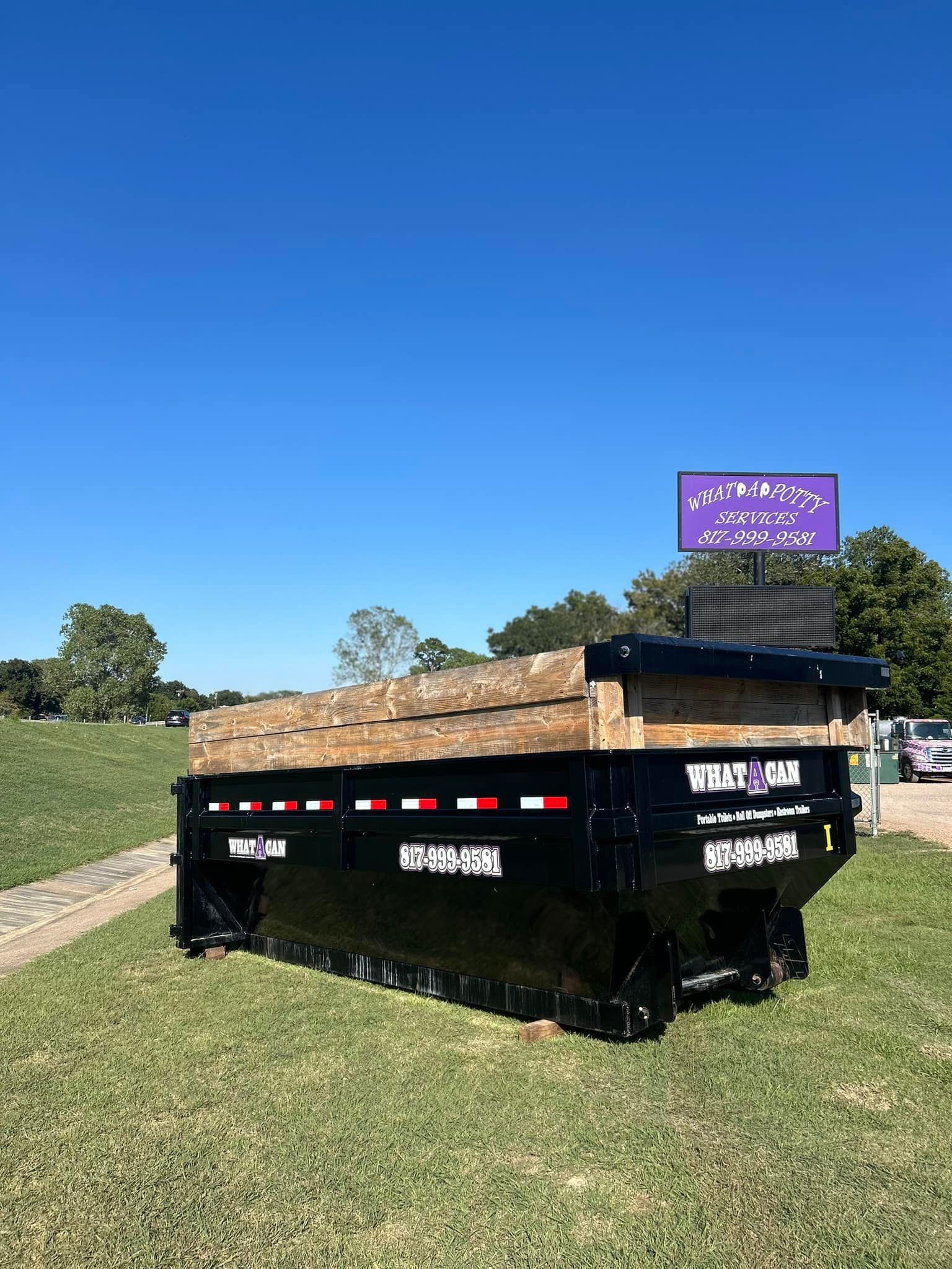 Black dumpster filled with wood sits on a grassy area, under a blue sky.