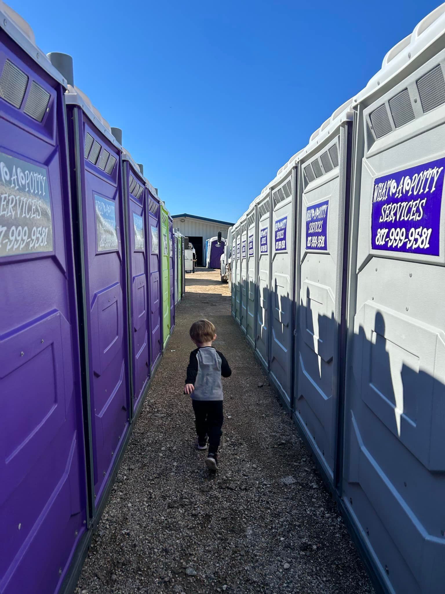 Child walking down an aisle of portable toilets. Purple and gray units line the path.