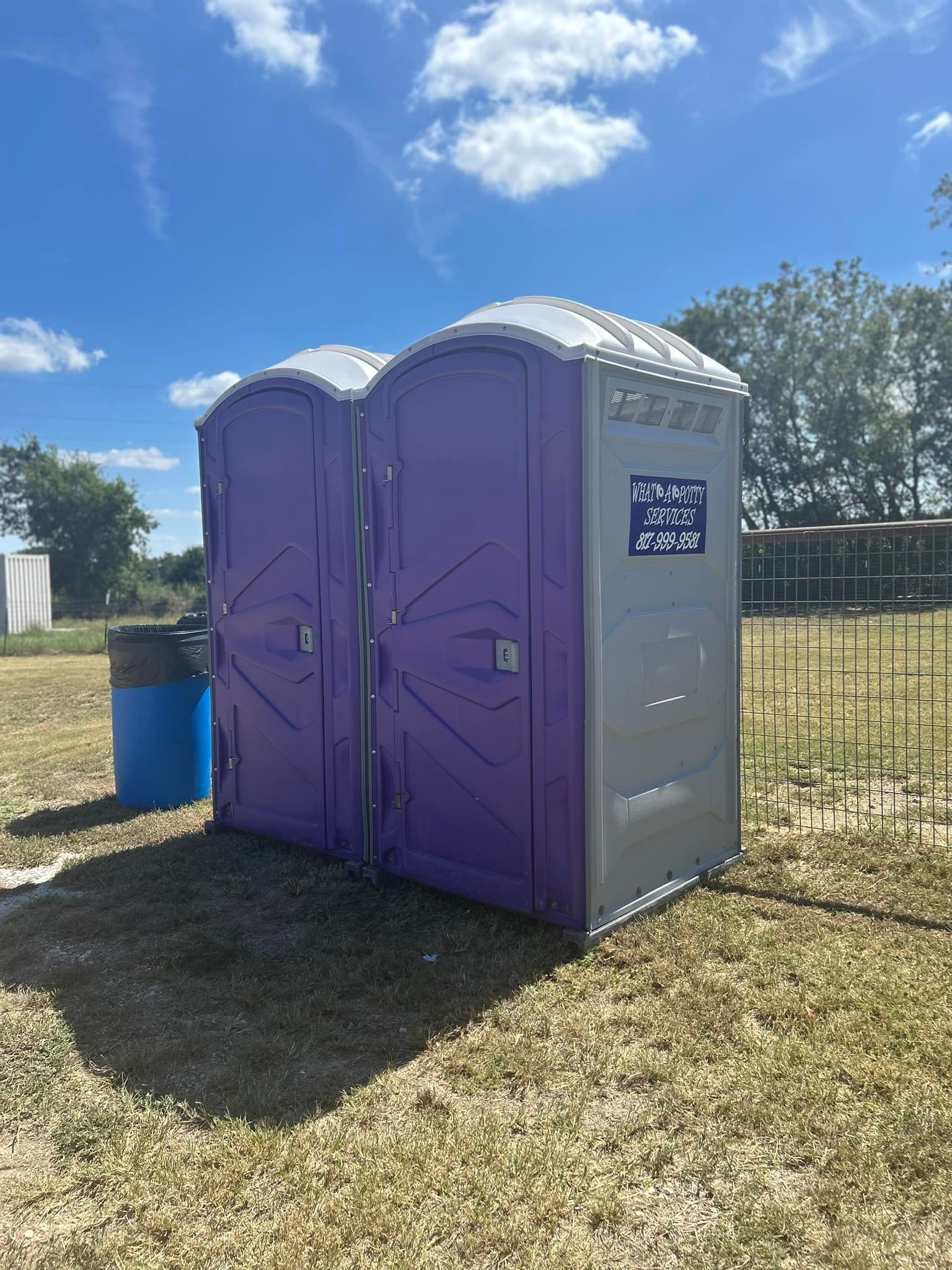 Two purple and gray portable toilets in a grassy outdoor setting under a blue sky.