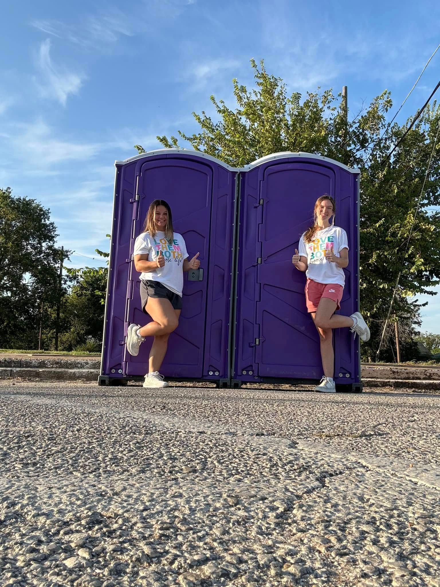 Two people stand by purple portable toilets outdoors.