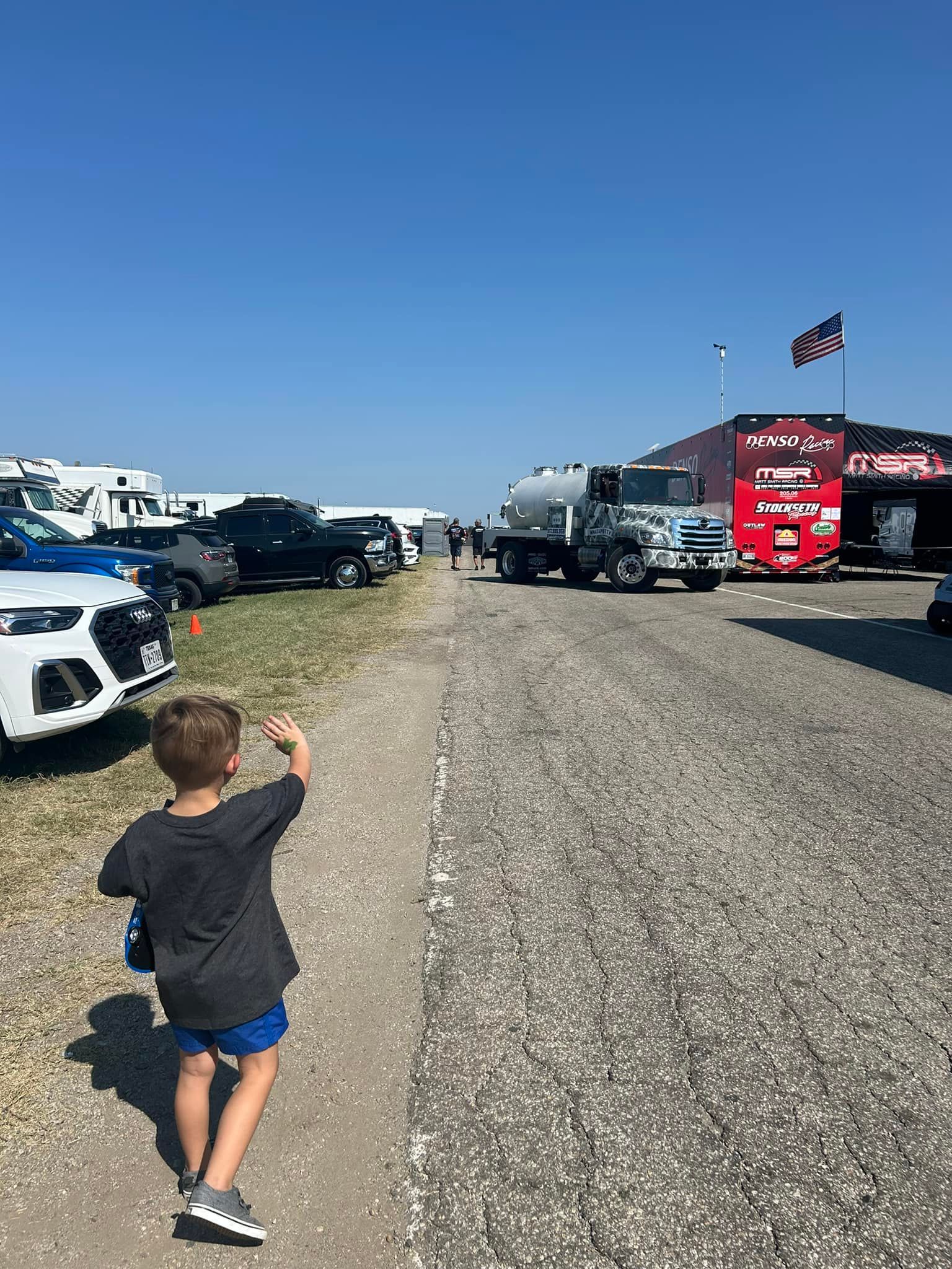 Boy points ahead on a gravel road, near parked vehicles and a large truck, under a blue sky.