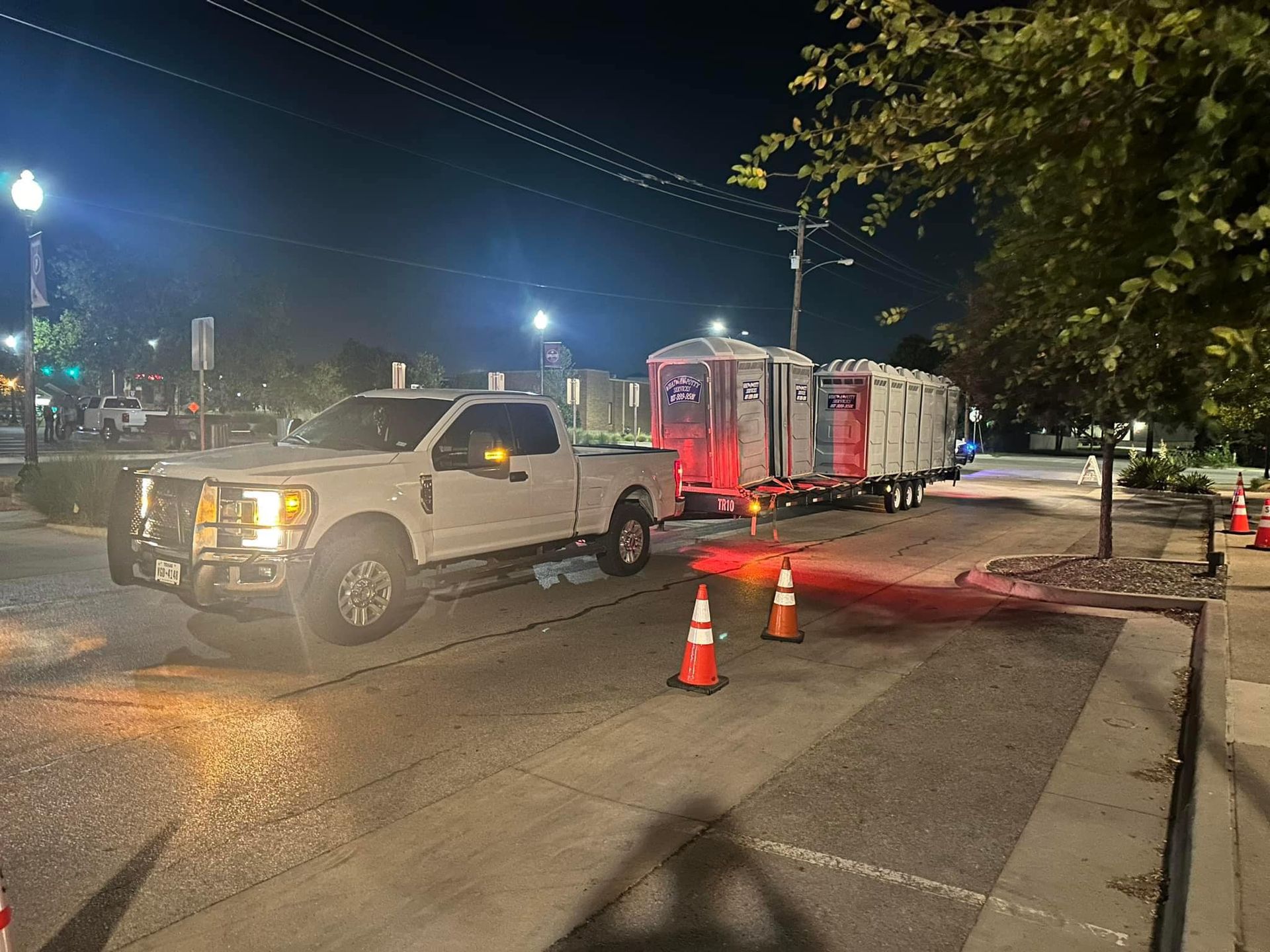 White truck pulling multiple trailers at night, with streetlights and orange cones.