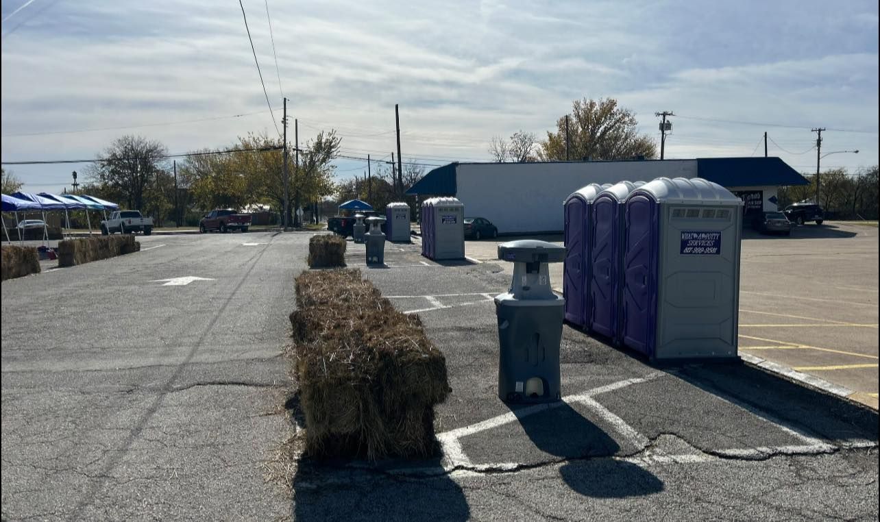 A row of portable toilets and hay bales on a parking lot, under a blue sky.