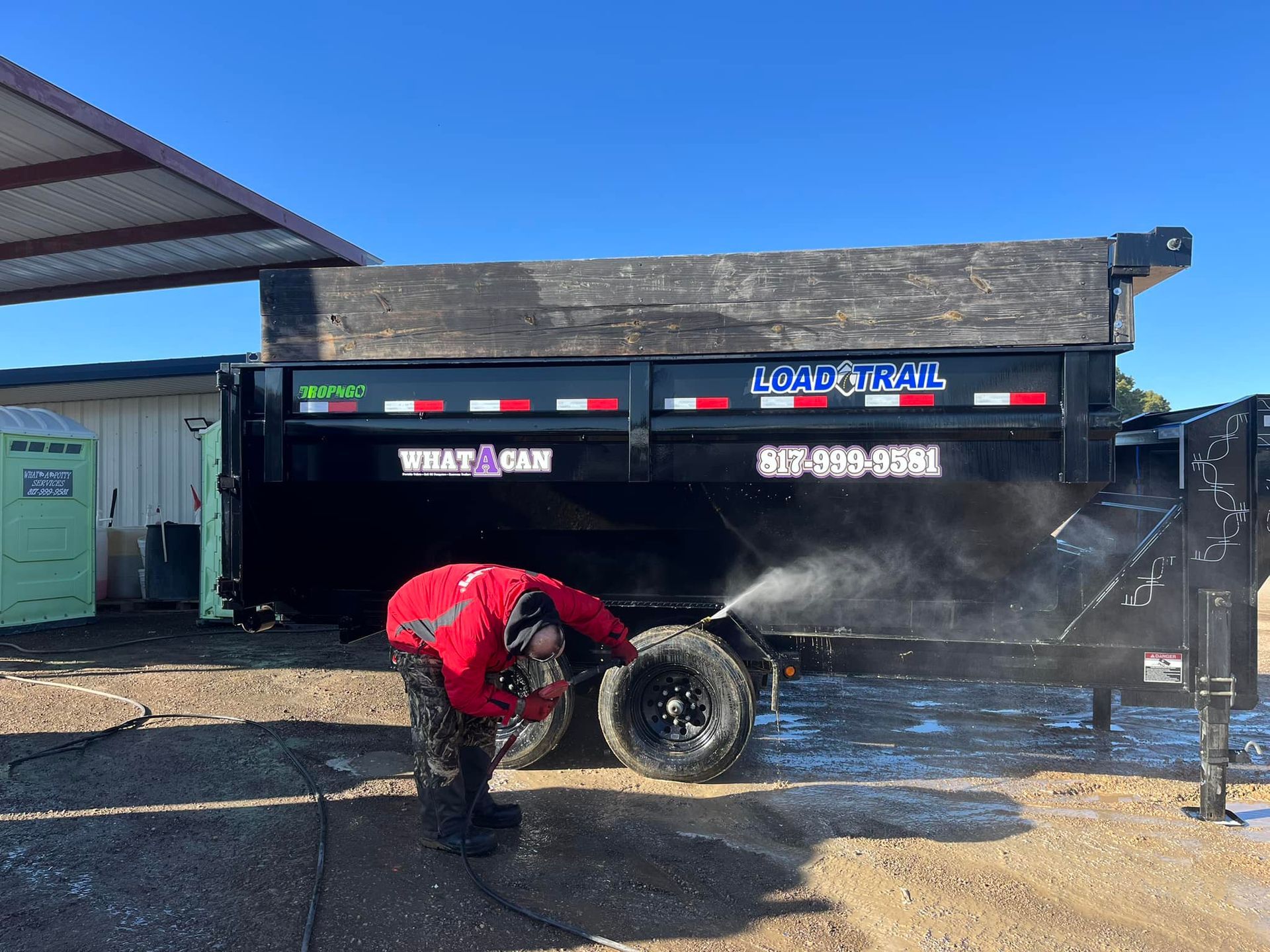 Person power washing the tire of a black Load Trail trailer outside on a sunny day.