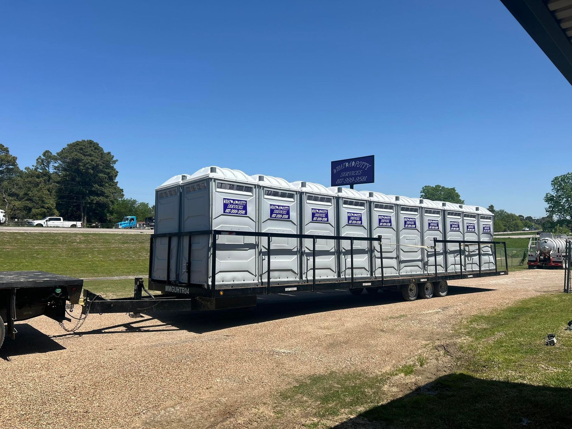 Portable toilets loaded on a trailer, parked on gravel in front of a blue sky.
