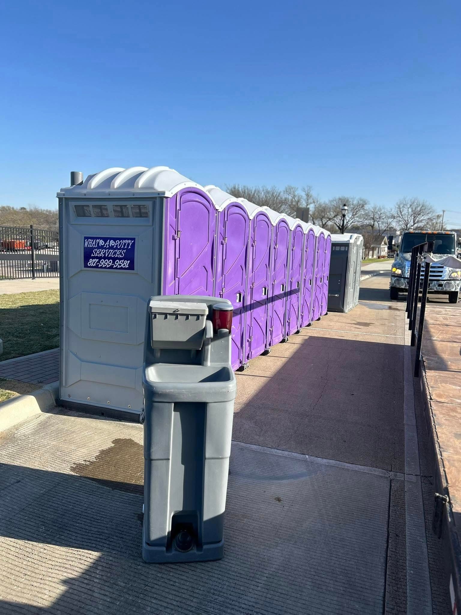 Row of portable toilets, some purple, on a paved surface next to a handwashing station under a blue sky.