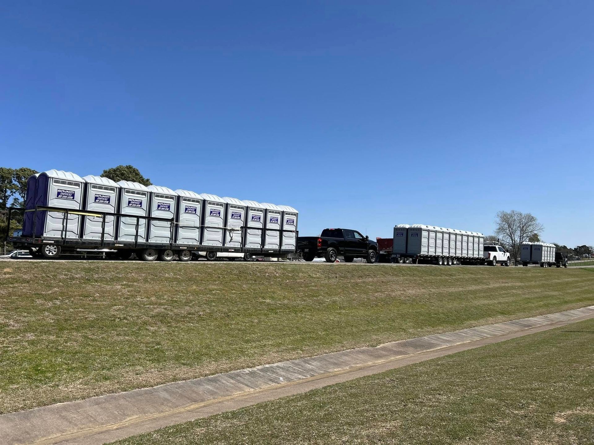 Two semi-trucks with trailers loaded with portable toilets on a grassy area under a blue sky.