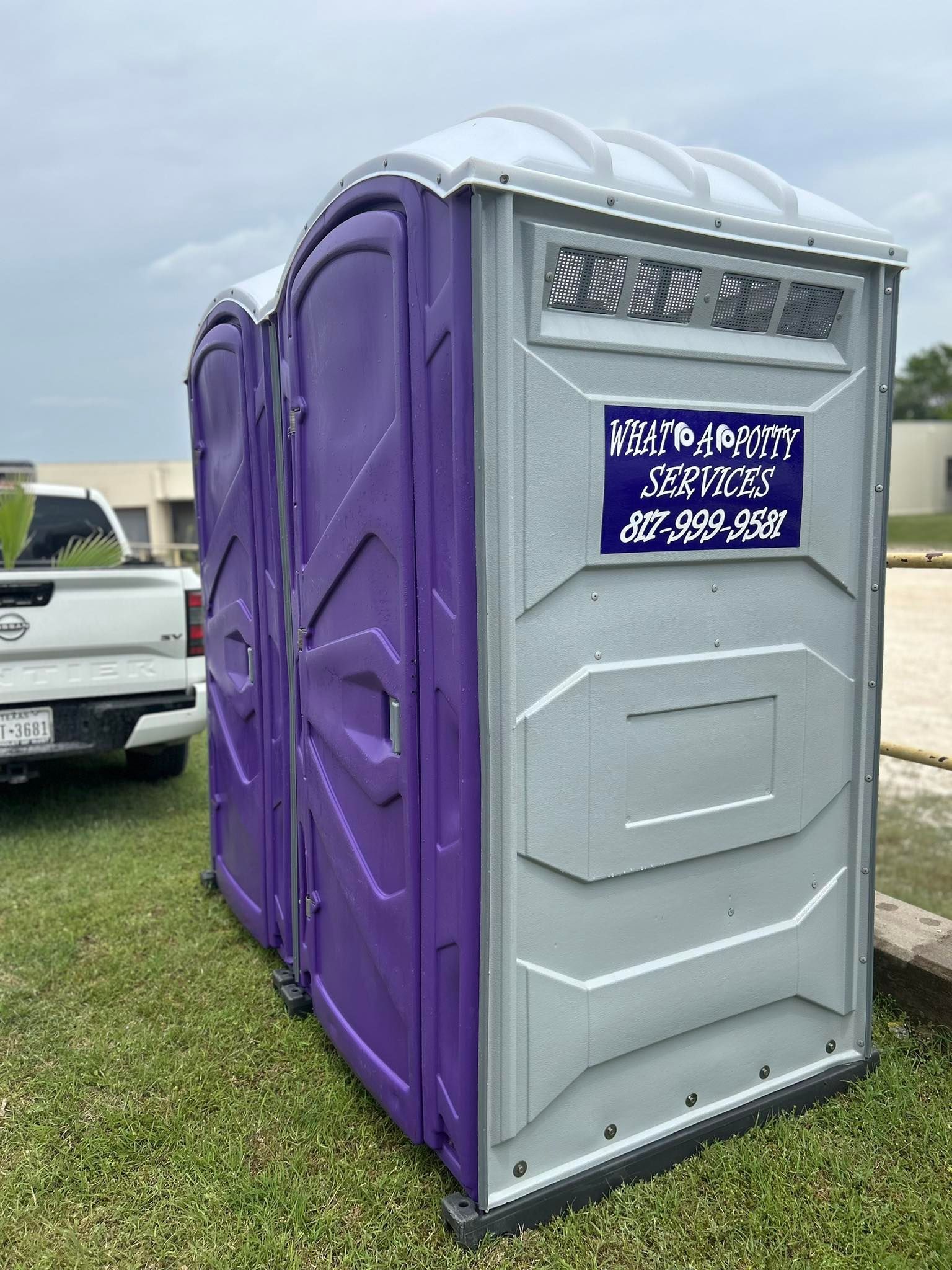 Three portable toilets on grass, one purple and two gray, under a cloudy sky.