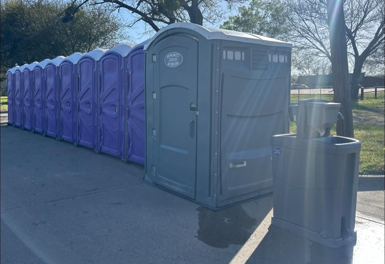 Row of purple and grey portable toilets on a concrete pad with a trash can in front.