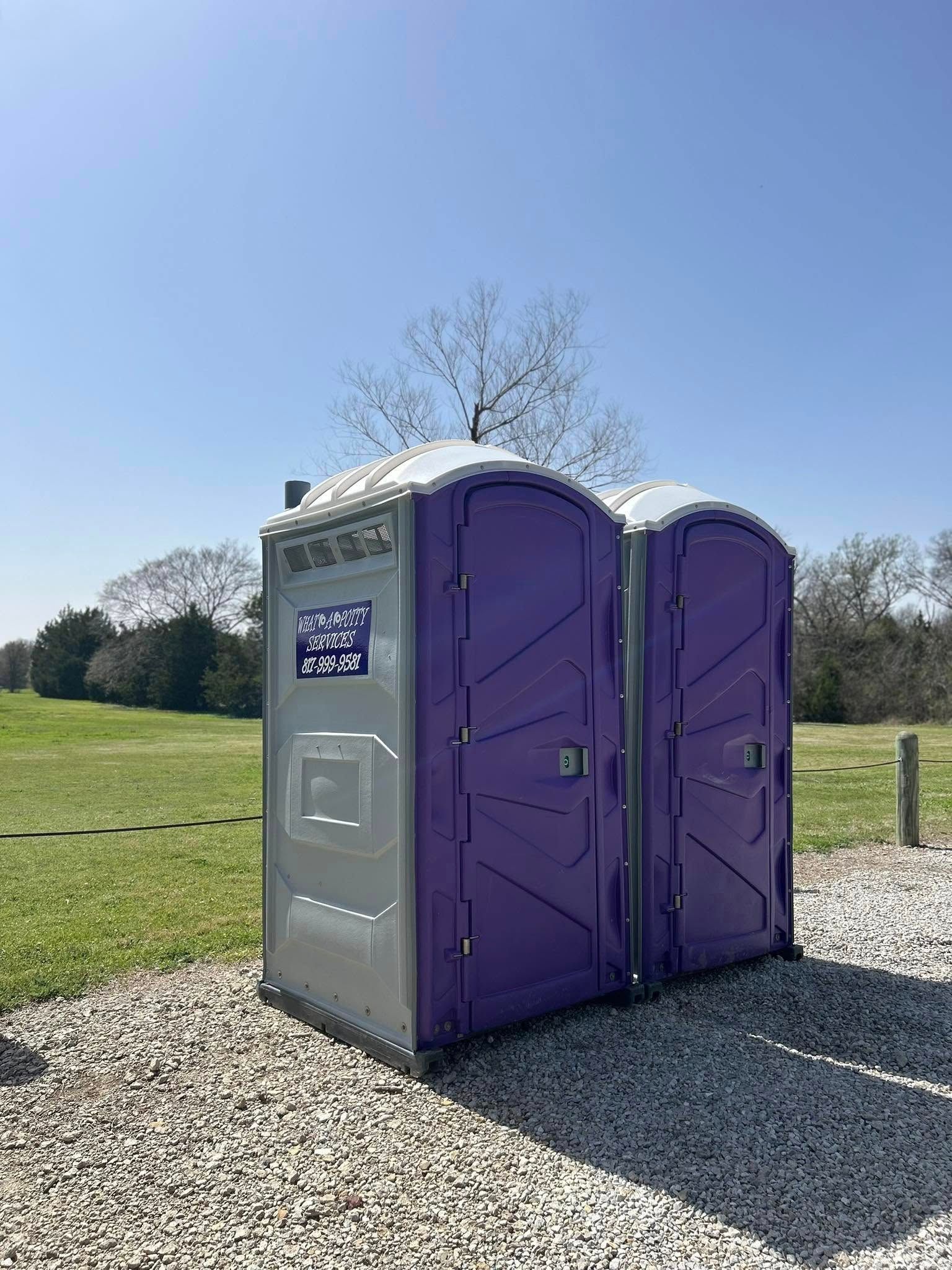 Two purple and gray portable toilets on gravel, outdoors with a tree and blue sky.
