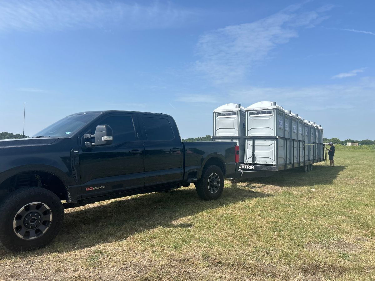 Black truck towing a trailer loaded with portable toilets on a grassy field under a blue sky.