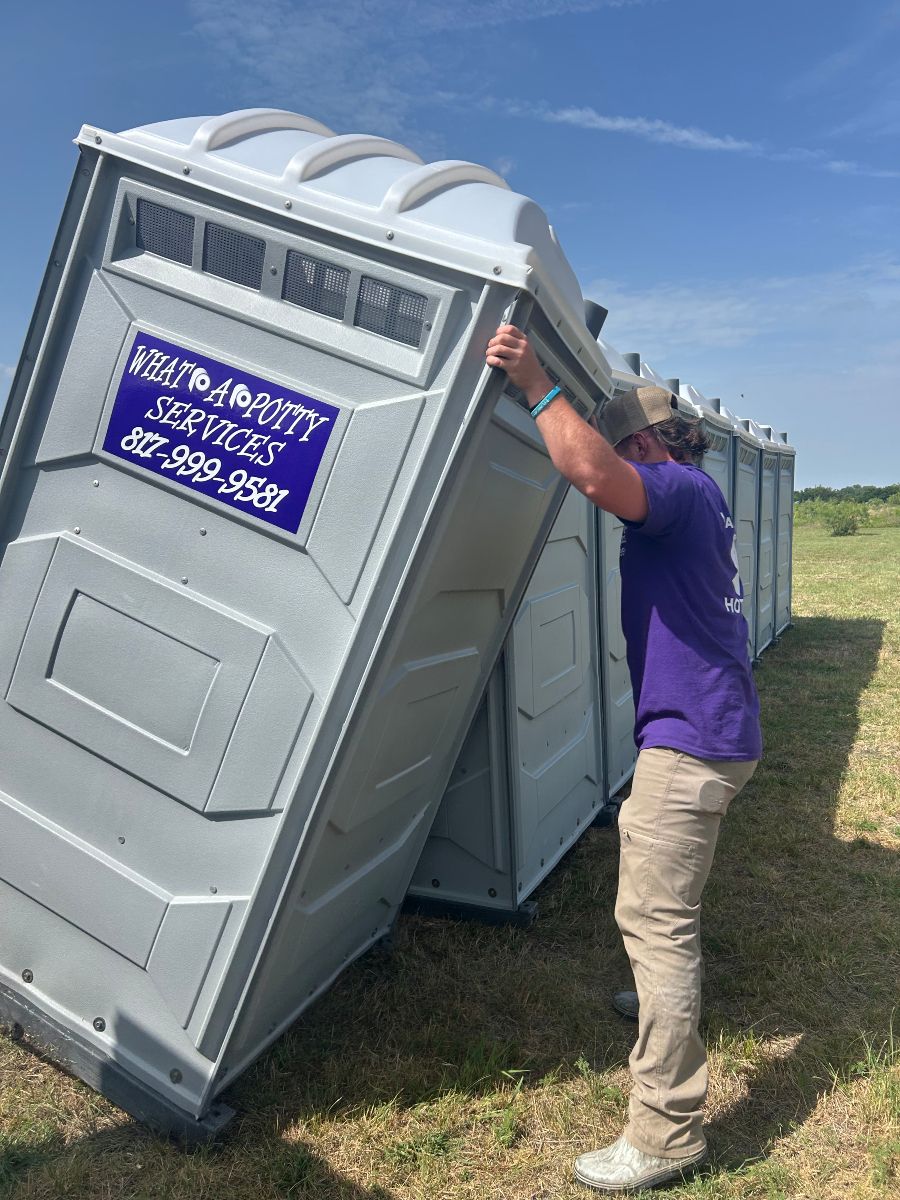 Man pushing a gray portable toilet. Others line up on a grassy field under a blue sky.