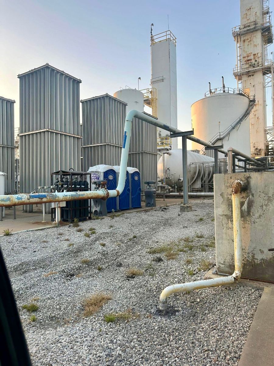 Industrial facility with tanks and piping against a pale blue sky. Ground covered in gravel with some weeds.