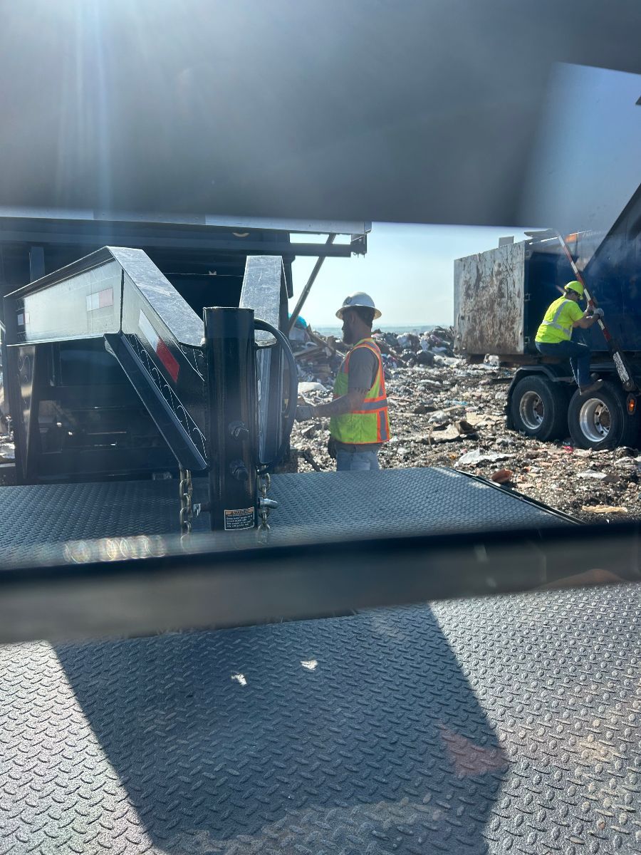Construction workers near a dump truck, wearing hard hats and vests, with waste in the background on a sunny day.