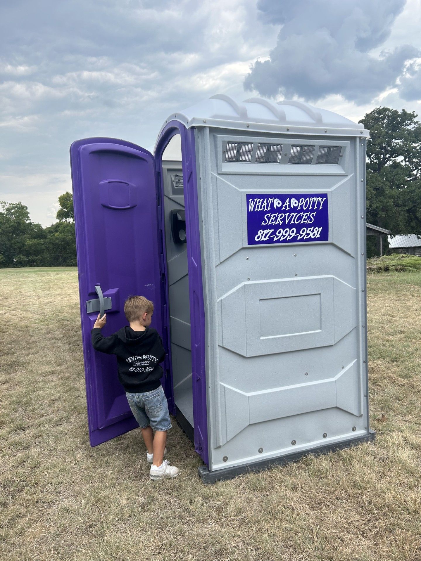 Child standing in the doorway of a portable toilet with a purple door, outdoors.