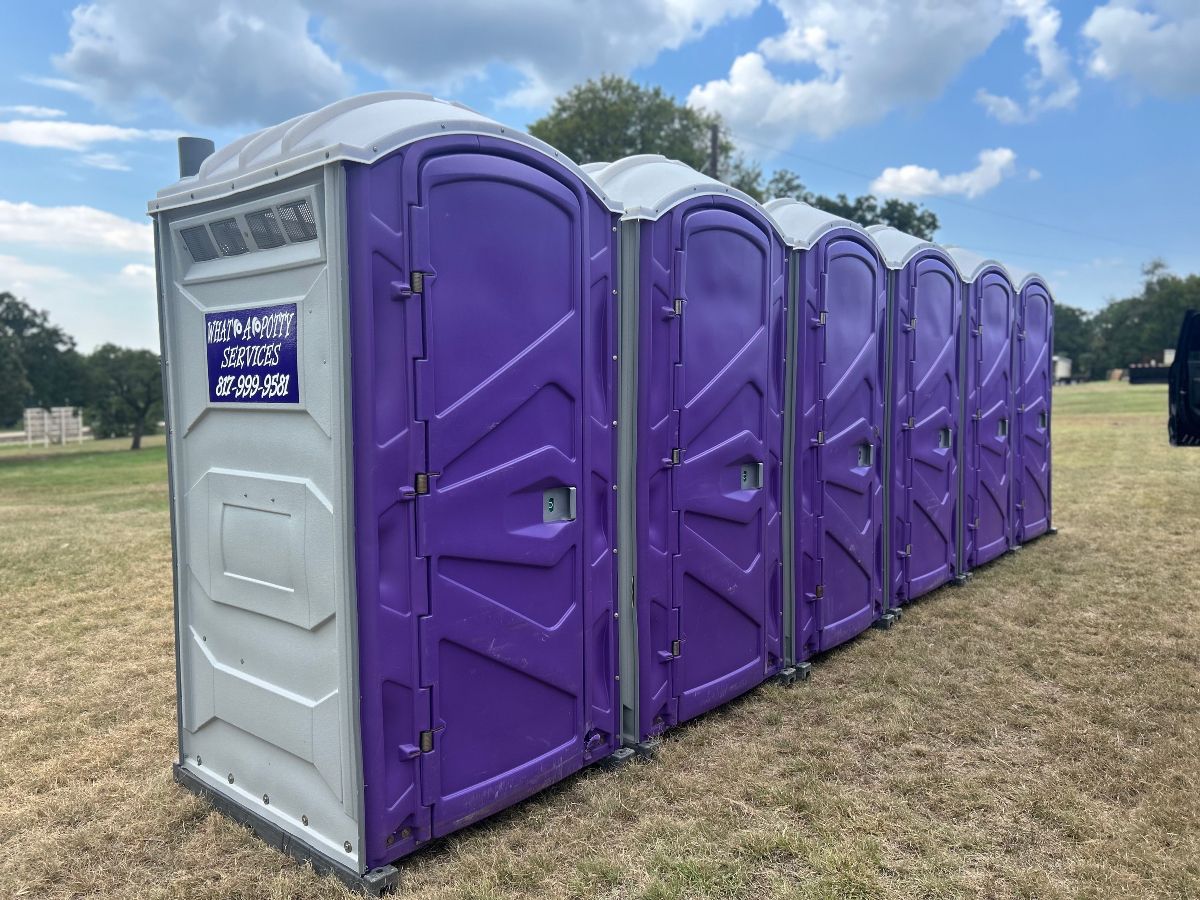 Row of purple and gray portable toilets outdoors on a grassy field under a cloudy sky.