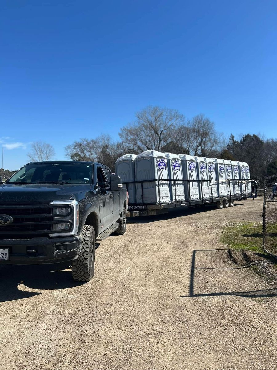 Black truck towing a trailer loaded with portable toilets on a dirt road under a blue sky.