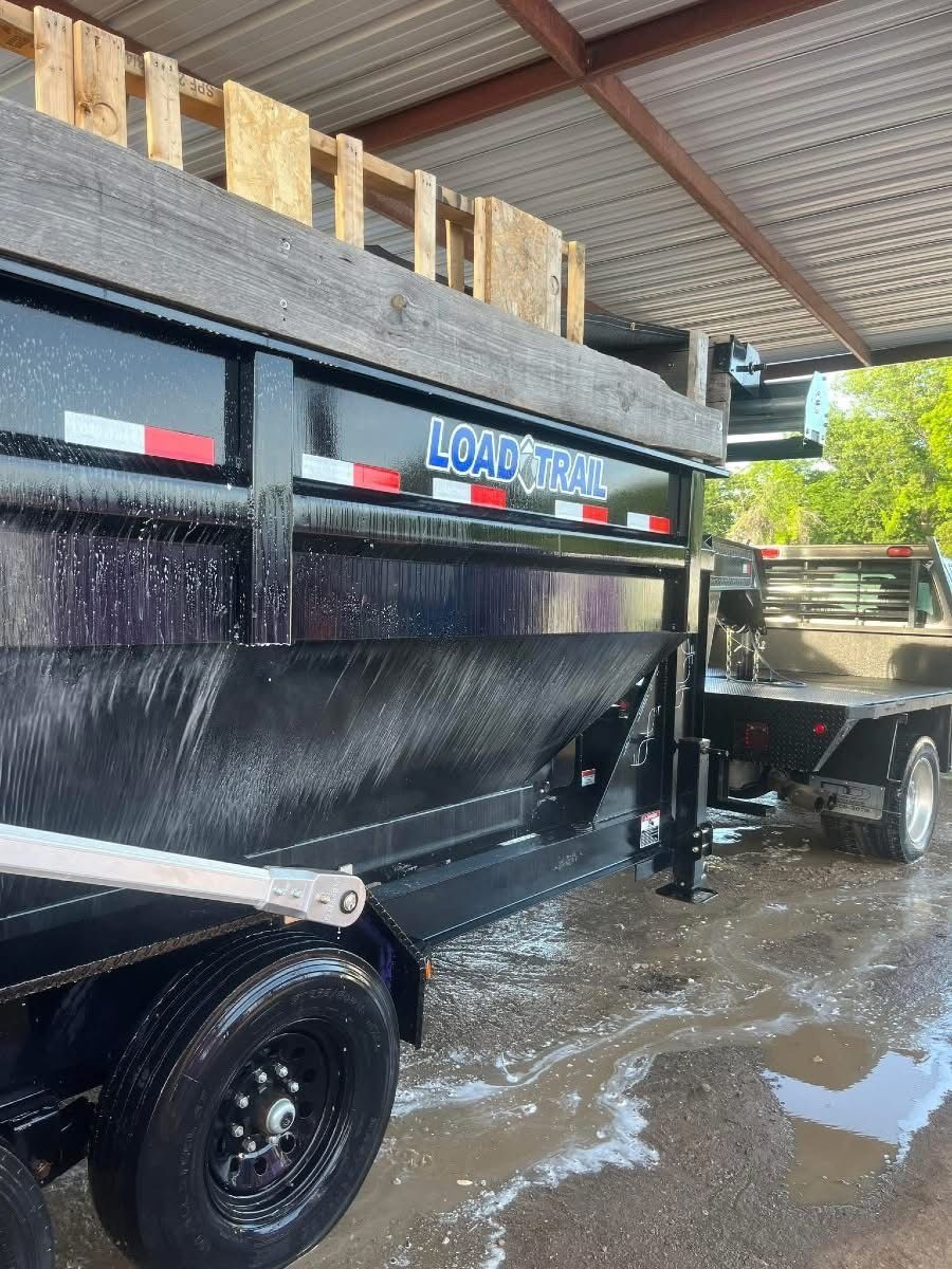 Black dump trailer attached to a pickup truck, under a metal roof, being washed.
