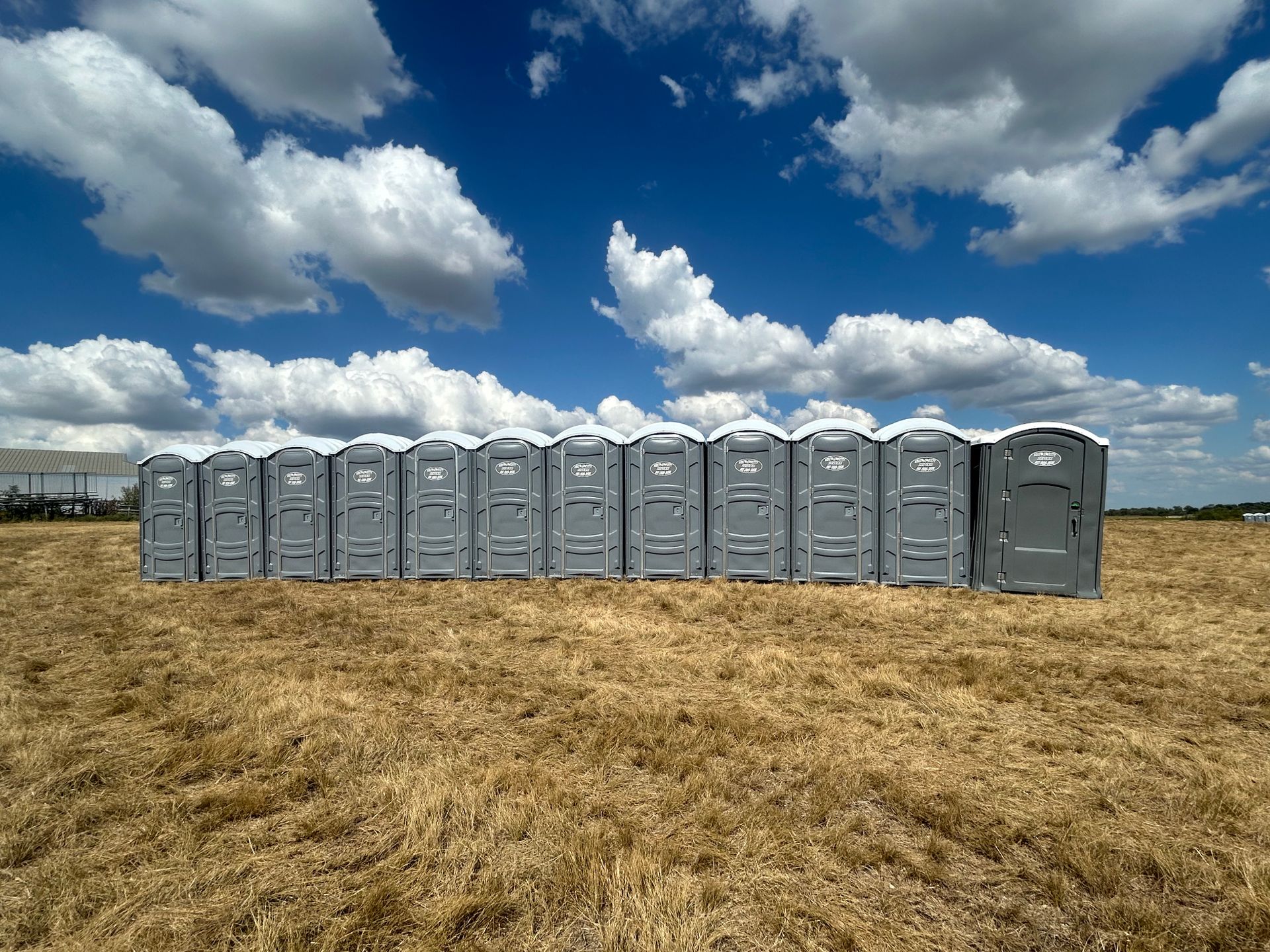 A row of gray portable toilets in a harvested field under a blue sky with fluffy clouds.