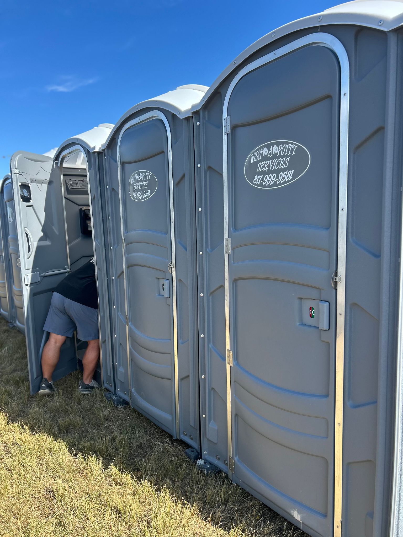 A row of gray portable toilets with a person inside one; outdoor setting.