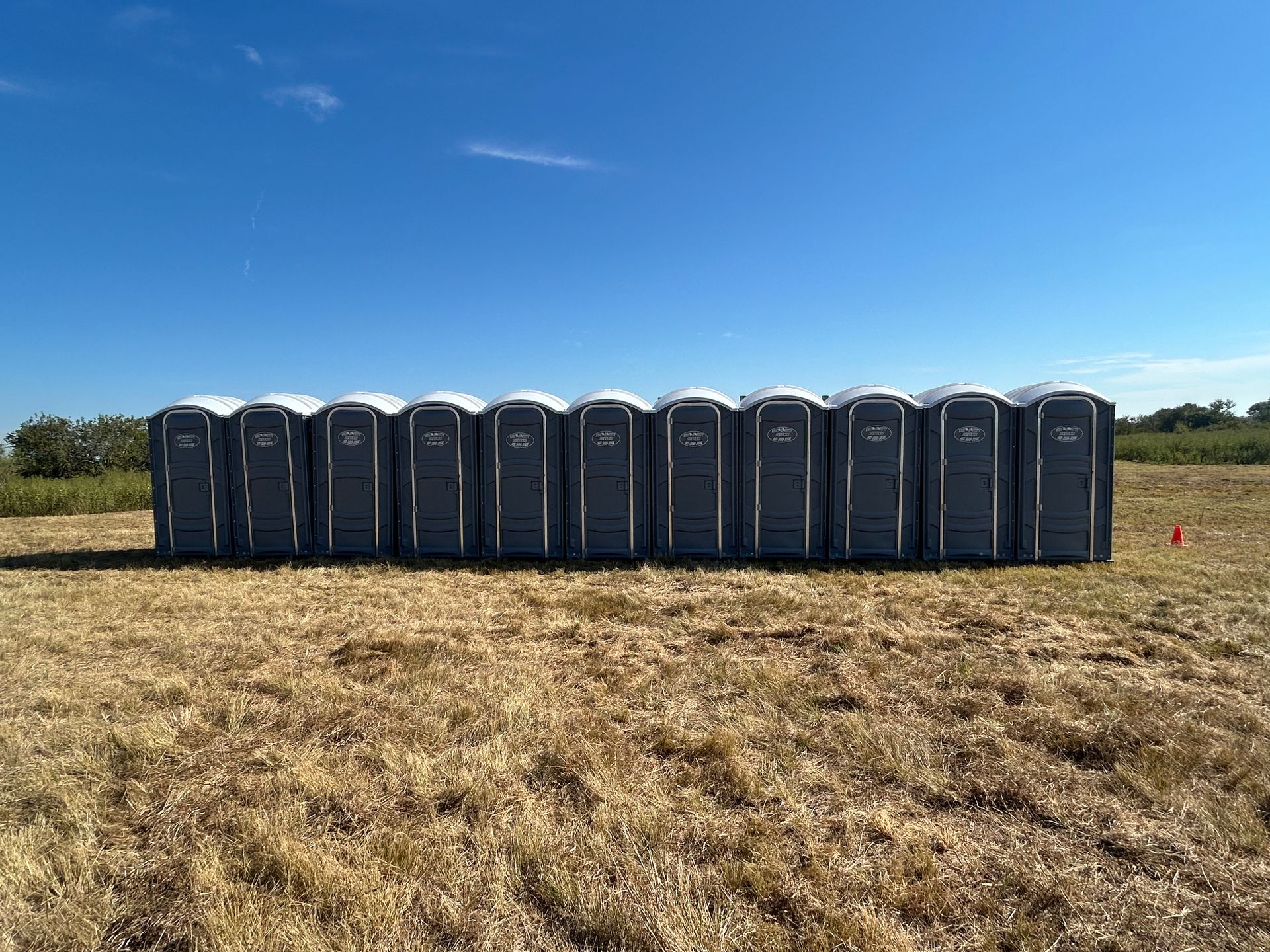 Row of gray portable toilets on a dry grassy field under a clear blue sky.