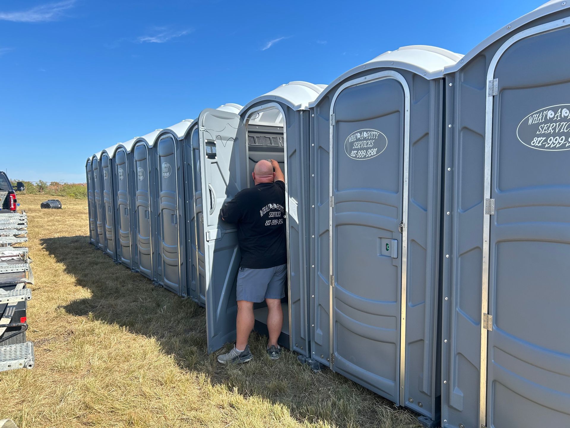 Row of gray portable toilets, one door open; a person is entering one. Outdoors, sunny.