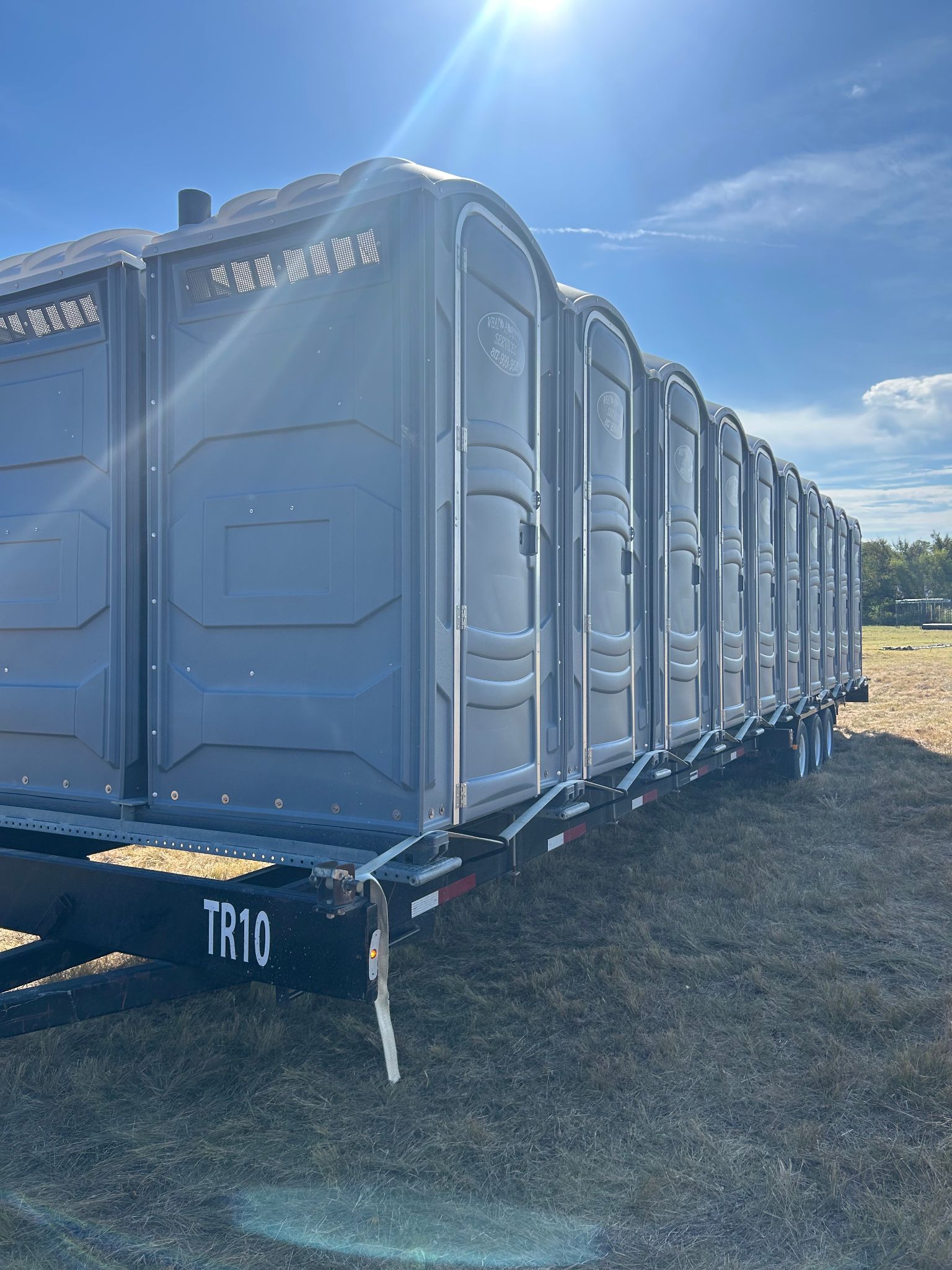 Row of gray portable toilets on a trailer, under a bright, sunny sky.