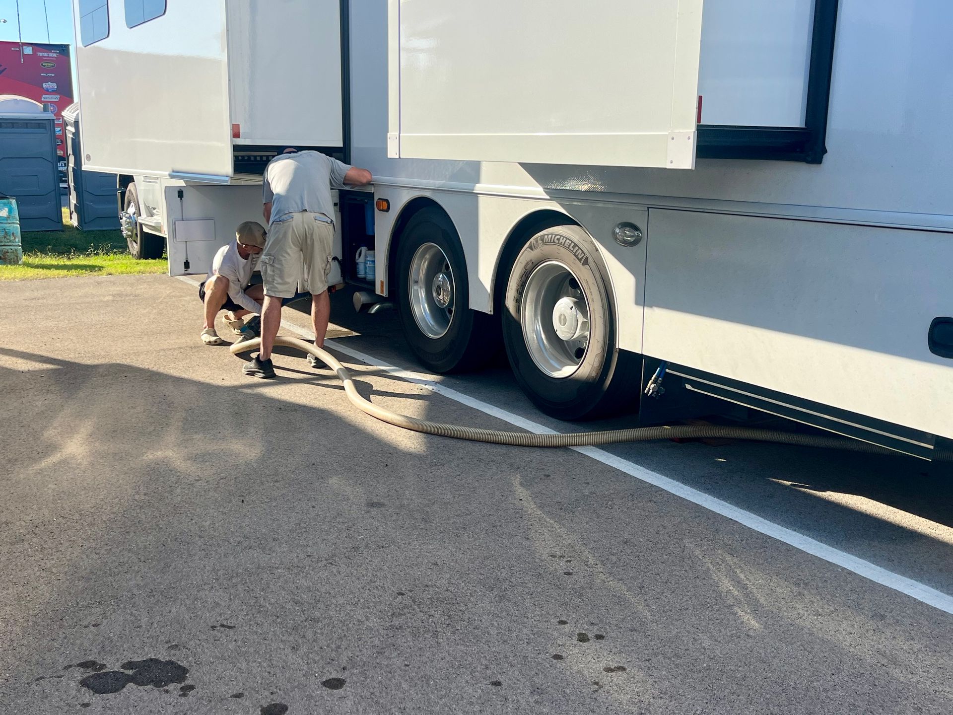 Two people attaching a large, thick hose to the side of a white truck on a paved surface.