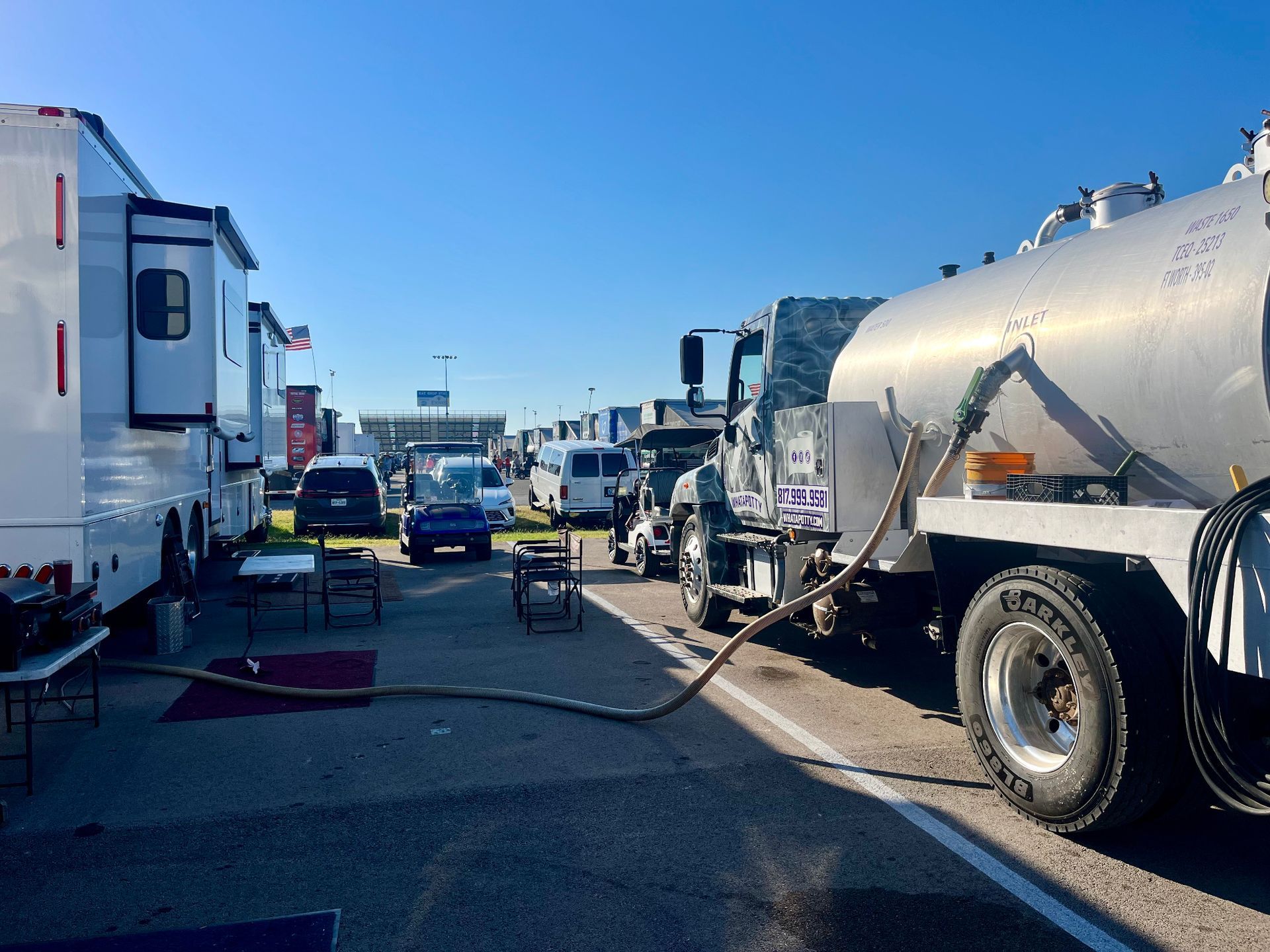A tanker truck fueling RVs parked on asphalt; several vehicles and tables are in the background under a blue sky.
