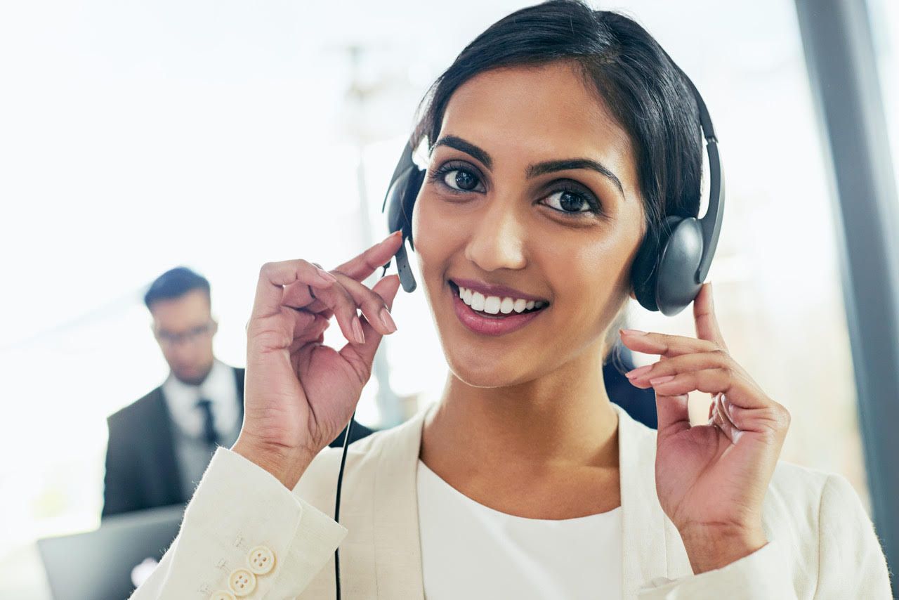 A professional wearing a headset smiles, hands touching the earpieces, in an office setting.