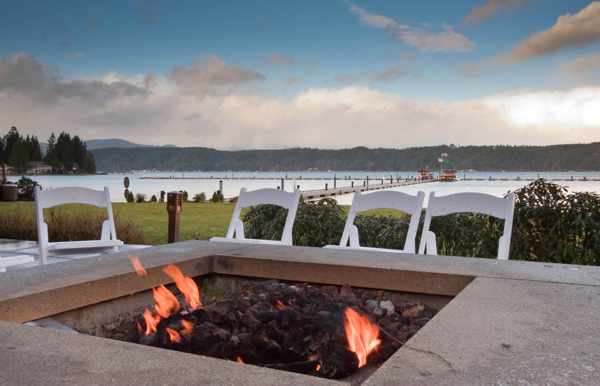 Fire pit by a lakeside patio with white chairs and people in the distance at sunset