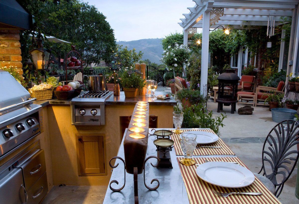 Outdoor patio dining area with grill, lit table, and lush plants overlooking a mountain view