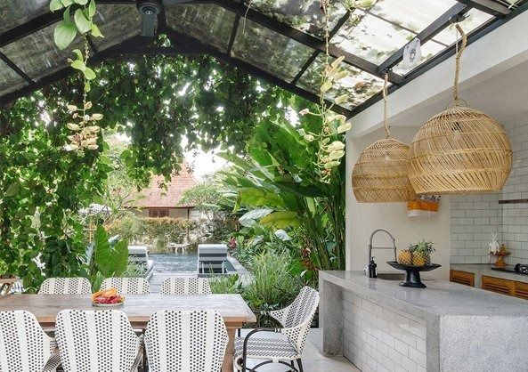 Open-air patio with white table setting, hanging wicker lights, and lush greenery under a glass roof