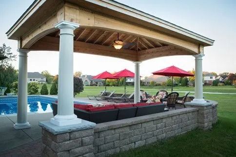 Covered backyard patio with red umbrella tables beside a pool and stone seating wall