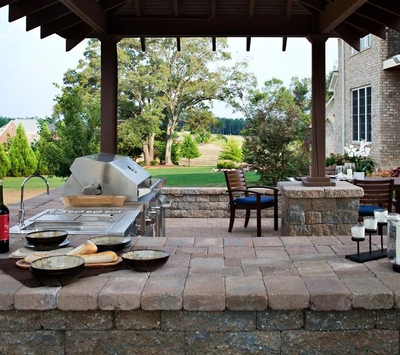 Covered patio with stone pavers, grill, table and chairs beside a garden and house exterior.