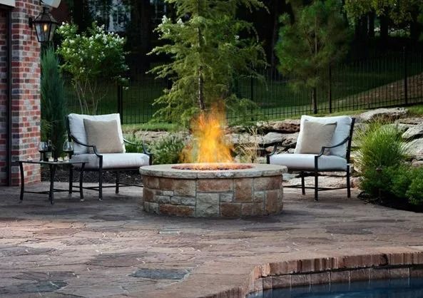 Backyard patio with two cushioned chairs around a lit stone fire pit beside a pool